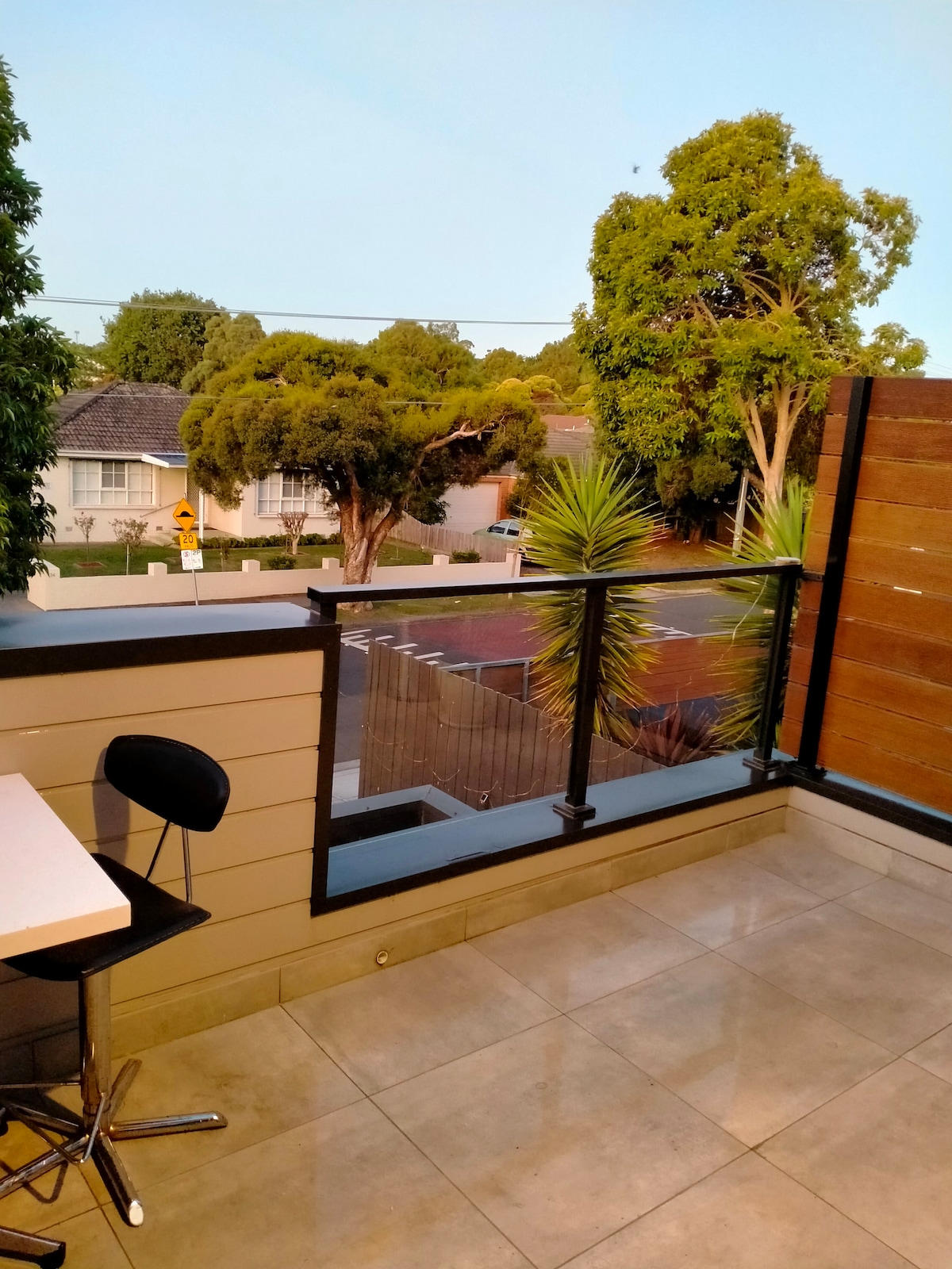 A balcony space is shown with modern gray tile flooring. A small table and chair set are positioned to one side, overlooking a view of trees and a neighborhood. The railing is a mix of glass and wood, providing a sense of openness.