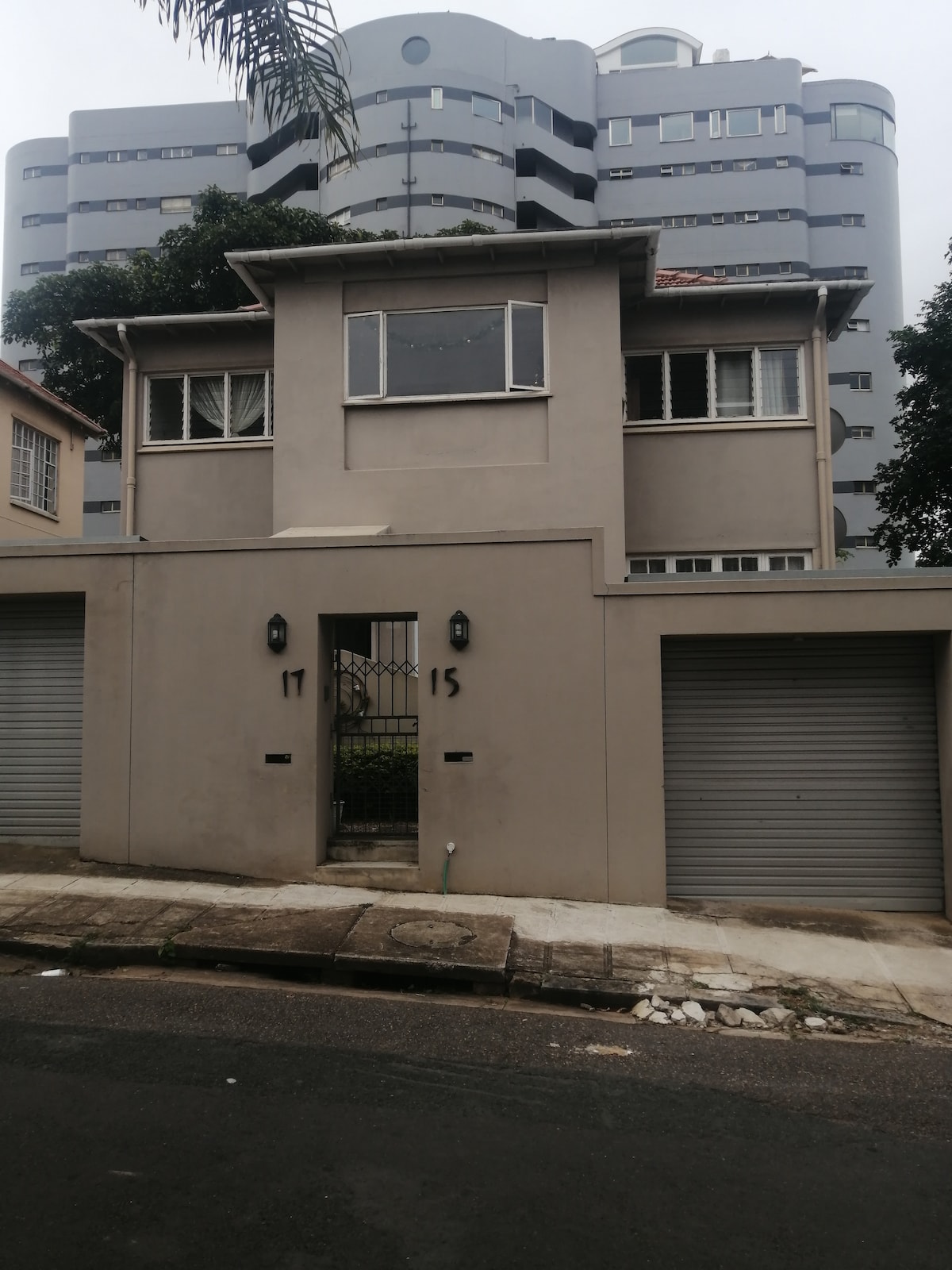 The exterior of a two-story building is visible, featuring a neutral-toned facade. Large windows provide natural light to the upper level, while two roll-up garage doors are situated on either side. A gated entrance adds a layer of security, highlighting the quiet street setting.