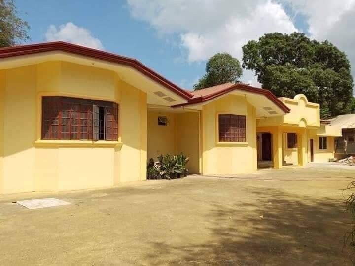 The exterior of a bright yellow structure is shown, featuring a red roof and multiple windows. Surrounding greenery creates a peaceful atmosphere. A spacious driveway is visible, leading to the entrance of the property.