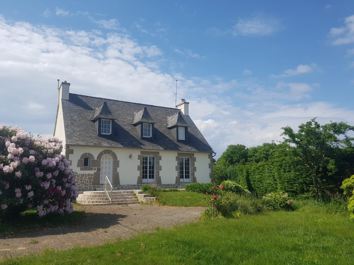 A charming house is set against a clear blue sky, surrounded by greenery and colorful flowering shrubs. The exterior features a slate roof and front steps leading to arched entryways. A spacious lawn with landscaped areas enhances the tranquil setting.