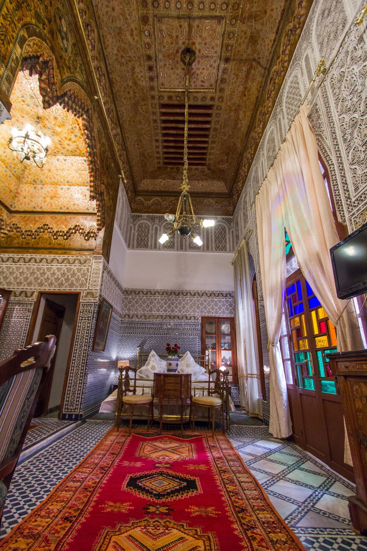 A bright dining area showcases an ornate ceiling with intricate patterns and a glass chandelier. Delicate wooden furniture surrounds a round table, set for a meal. Colorful stained glass windows add vibrancy, while a traditional patterned rug complements the tiled floor.