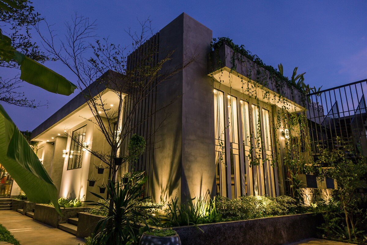 The exterior of the studio is presented at dusk, featuring a modern concrete façade complemented by wooden slats. Soft lights illuminate the structure and surrounding greenery, creating a serene ambiance. Lush plants frame the entrance, enhancing the welcoming atmosphere.