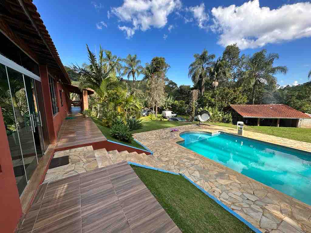 A spacious outdoor area features a clear swimming pool surrounded by lush greenery. Pathways made of stone and tile lead to a covered patio with large glass doors, providing a view of the scenic landscape and mature trees.