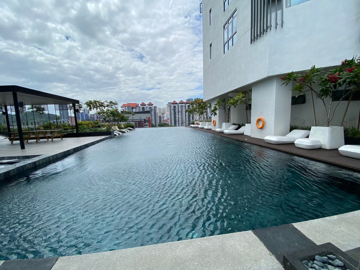 An inviting infinity pool sits against a modern building, surrounded by well-maintained greenery. Lounge chairs are positioned along the poolside, inviting relaxation. The serene water reflects the cloudy sky above, creating a tranquil atmosphere for enjoyment.