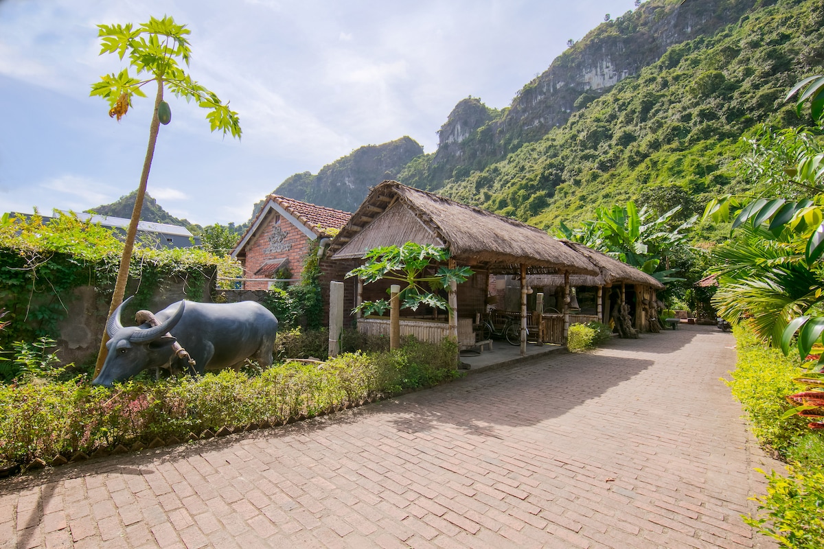 A traditional thatched-roof bungalow is situated alongside a winding brick pathway, surrounded by lush greenery and mountainous terrain. A decorative statue of a buffalo is positioned in the foreground, adding cultural charm to the inviting landscape.
