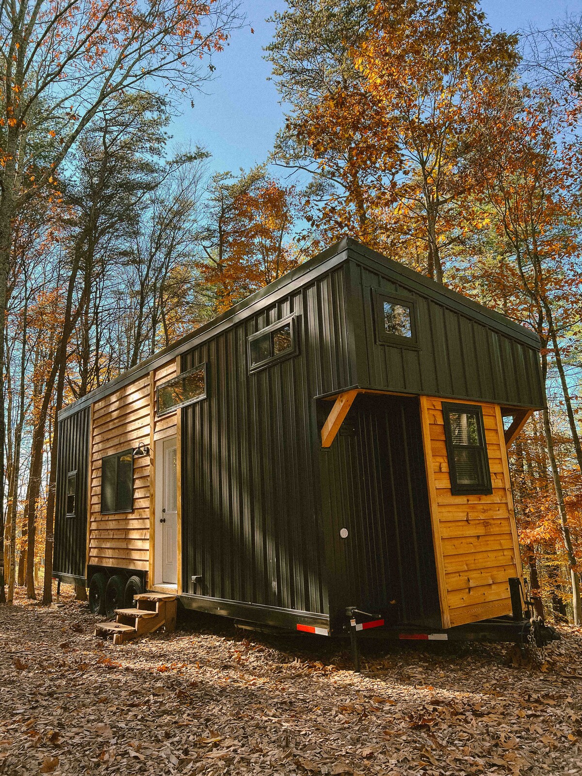 The tiny home is set in a forested area, featuring a mix of black and wood paneling. A small staircase leads to the front door, while large windows allow natural light to enhance the exterior design. Surrounding fall foliage creates a vibrant backdrop.
