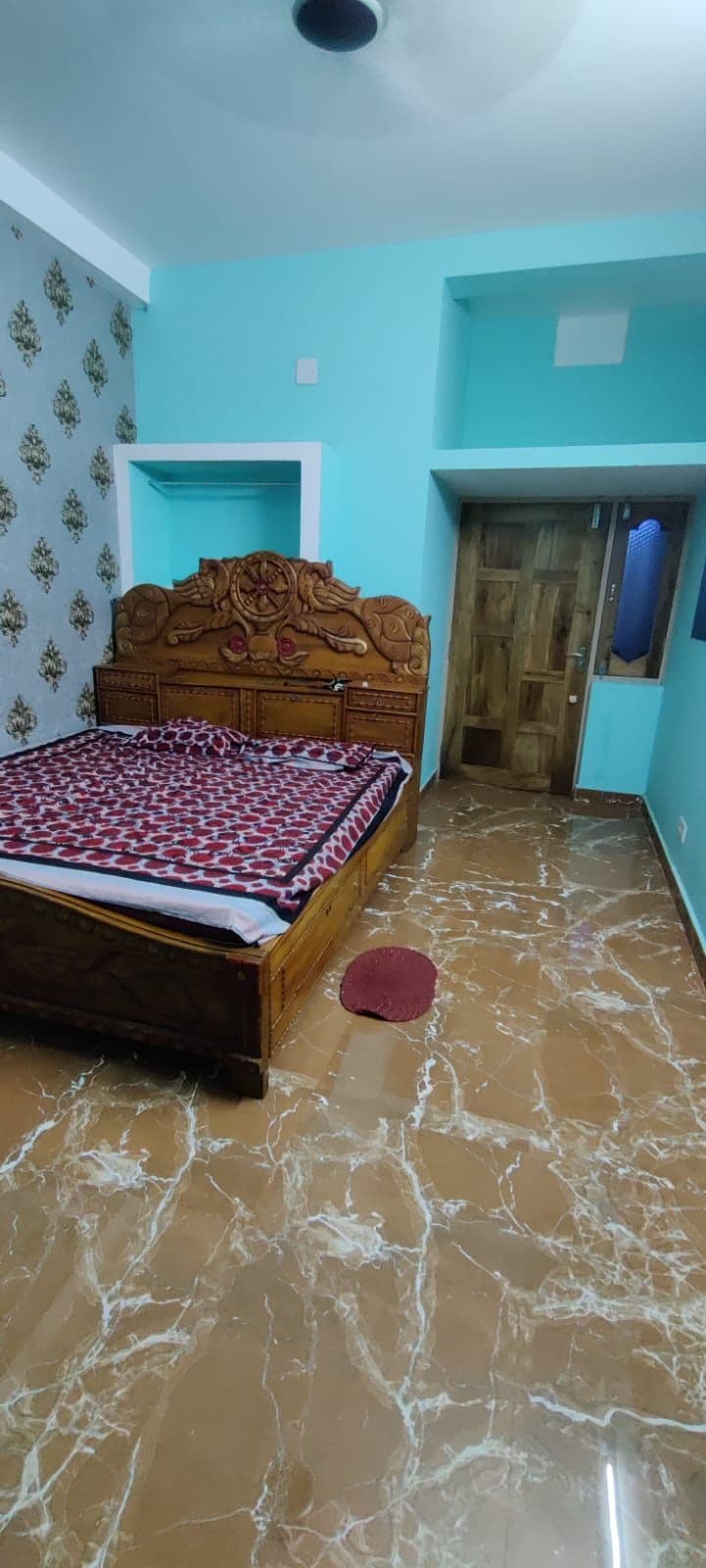 A bedroom is furnished with a decorative wooden bed featuring an intricately carved headboard. Colorful bedding covers the mattress, and the floor is finished with glossy tiles. A door leads to another room, while a wall is adorned with subtle patterned wallpaper in greenish tones.
