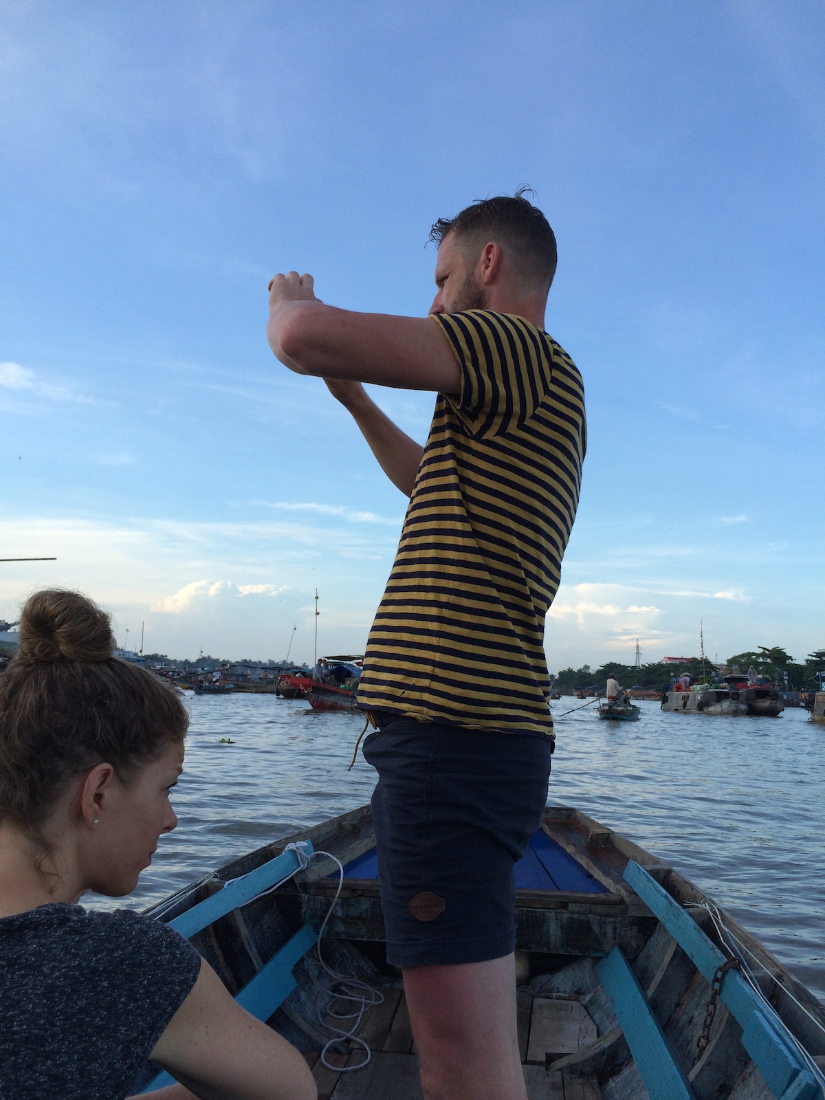 A view from a boat on the water, featuring two individuals. One person stands, taking a photo with a camera, while the other sits nearby. The sky is expansive with soft clouds, and various boats are visible on the water in the background.