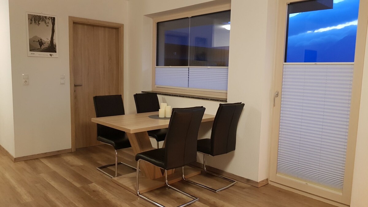 A dining area is presented with a wooden table surrounded by four black chairs. Light-colored walls and a large window provide natural light, while the view features the sky transitioning to twilight. A door leads outside, complementing the modern interior design.