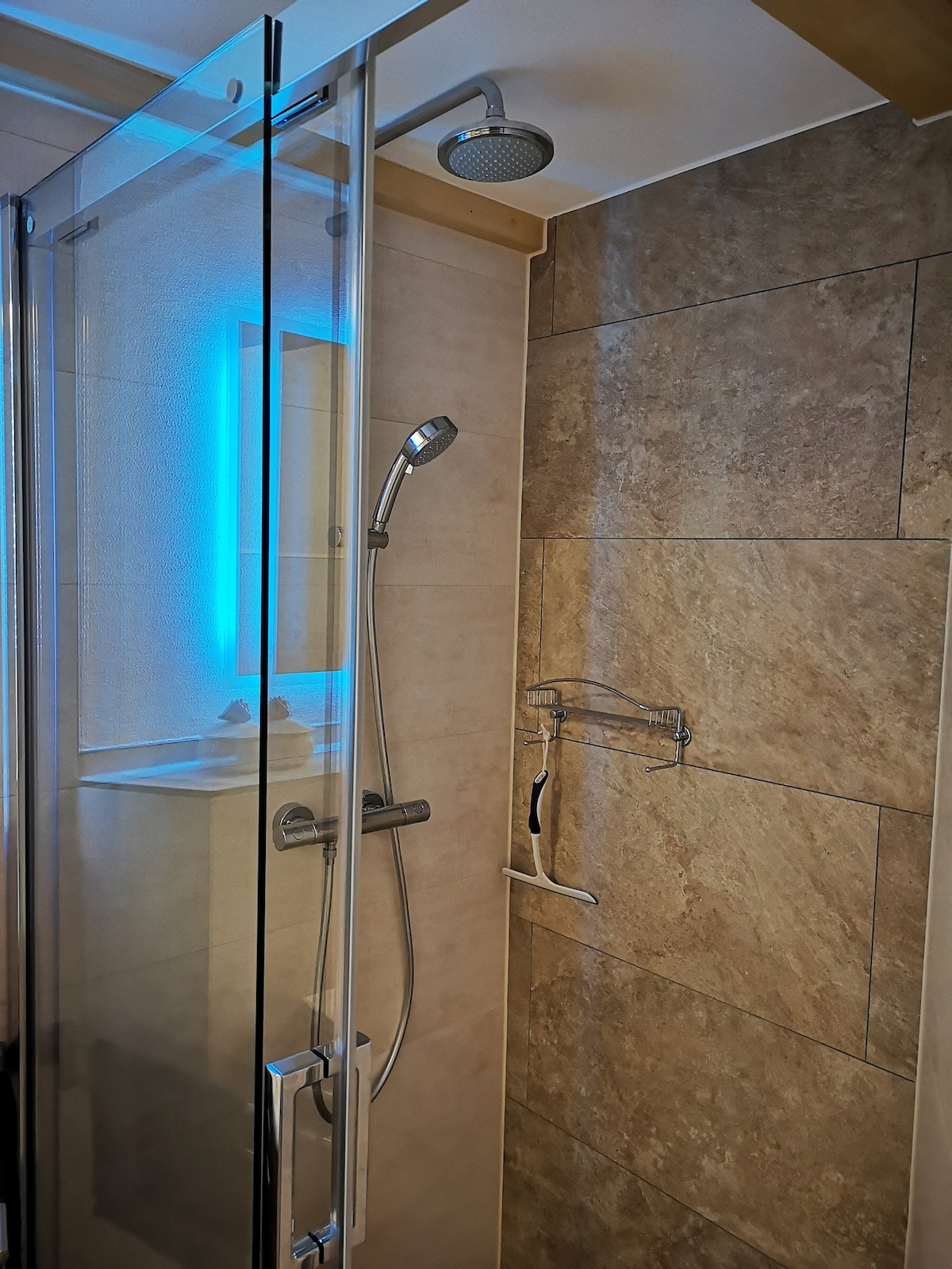 A modern walk-in shower features sleek glass panels and neutral stone tiles. An overhead rain shower head and a handheld shower head are visible. A backlit mirror provides adjustable color lighting, enhancing visibility in the space. A shower caddy is mounted on the wall for convenience.