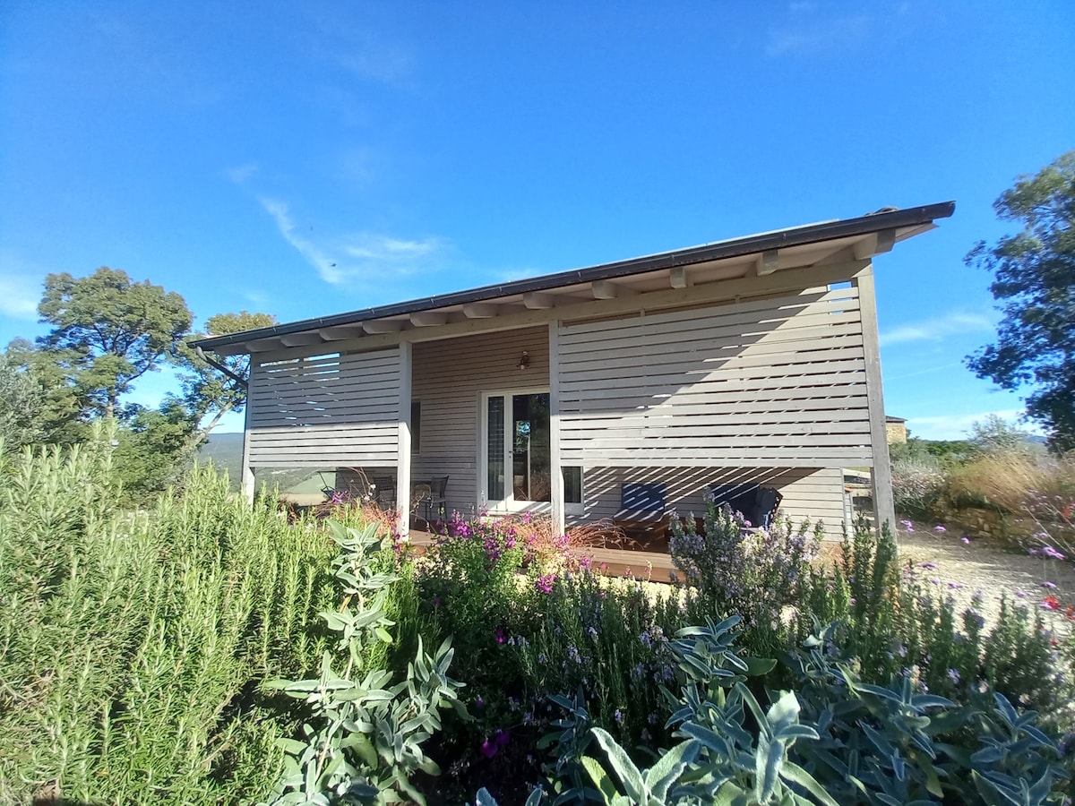 An eco-friendly house features a contemporary wooden façade with horizontal slats, allowing natural light while providing shade. Surrounding the exterior, colorful flowers and greenery enhance the serene rural setting. Large doors invite guests to experience the tranquil atmosphere within.