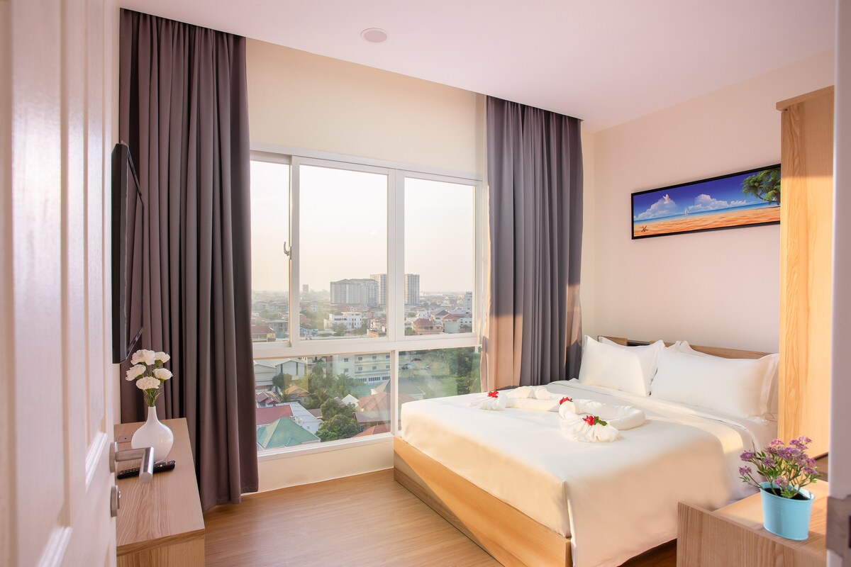 A bright bedroom features a spacious bed dressed in neutral linens, complemented by decorative flowers. Large windows provide a view of the surrounding area, with curtains adding a touch of elegance. A small table holds a potted plant, enhancing the room's inviting ambiance.