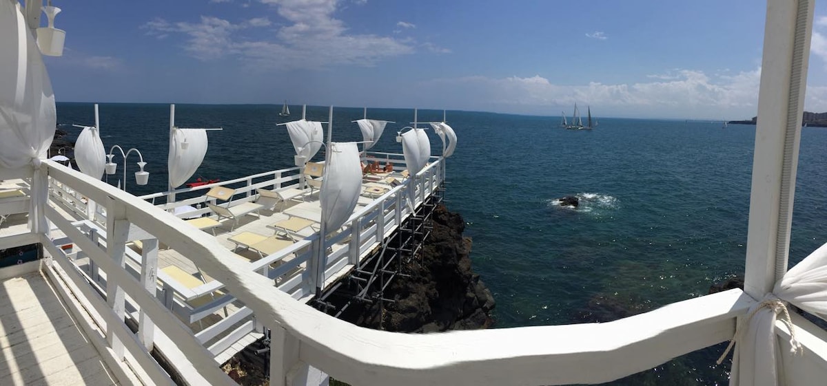 A spacious seaside deck extends over tranquil waters, with several sun loungers arranged for relaxation. Soft white sails gently billow in the breeze, providing shade. The horizon features distant boats, and rocky outcrops are visible nearby, enhancing the coastal atmosphere.