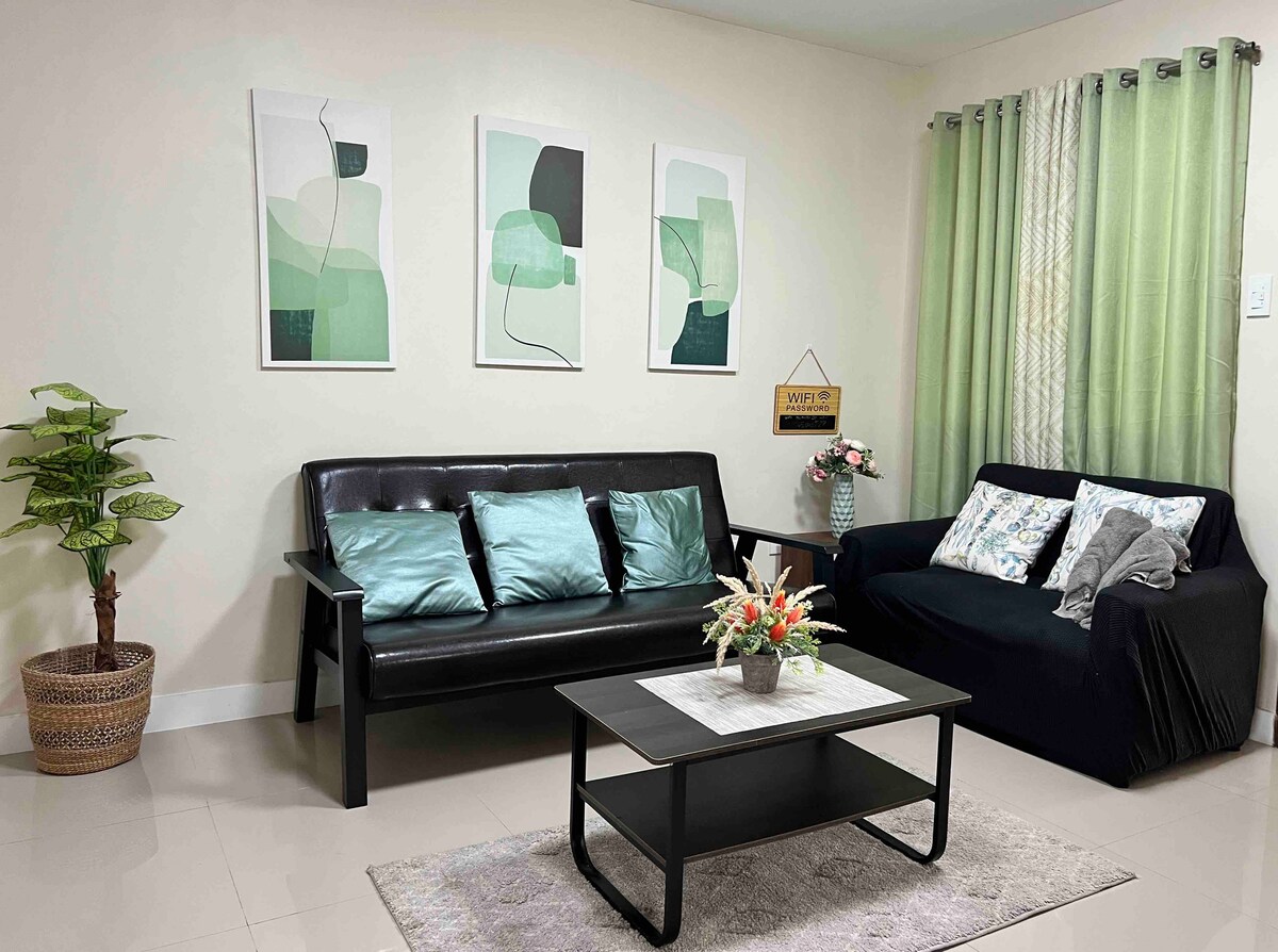 The living area features two sofas, one in black and another in dark fabric, complemented by decorative cushions. A coffee table is centered between them, and three abstract artworks adorn the wall above. A small plant adds a touch of greenery to the space.