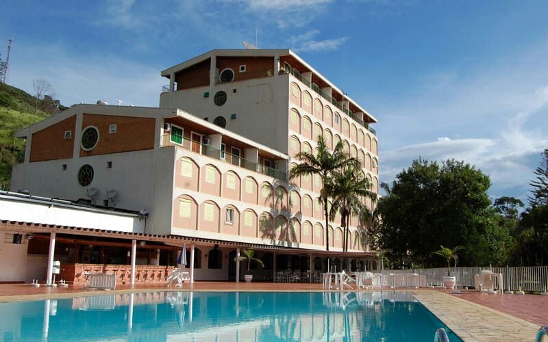 A multi-story building is seen beside a large outdoor pool, reflecting the blue sky. There are palm trees and open spaces around the pool area, contributing to a relaxed atmosphere. The architecture features rounded windows and beige-colored walls.
