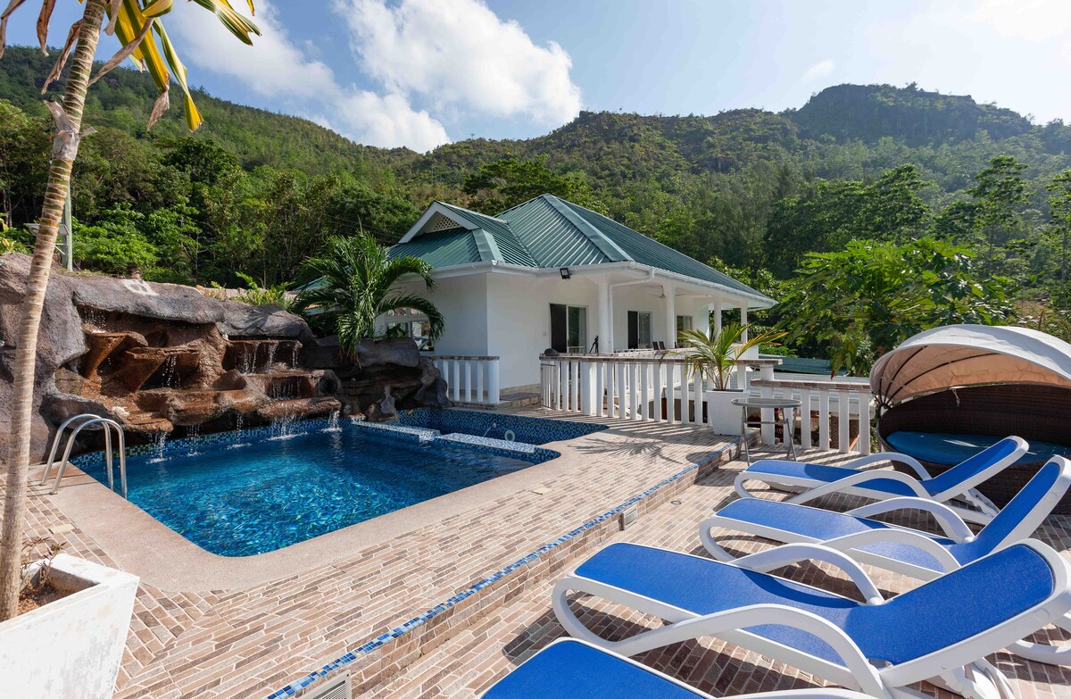 An inviting outdoor pool area is displayed with clear water, surrounded by lounge chairs. A beautiful house with a green roof and large windows sits nearby, framed by lush foliage and mountains in the background, enhancing the serene environment.