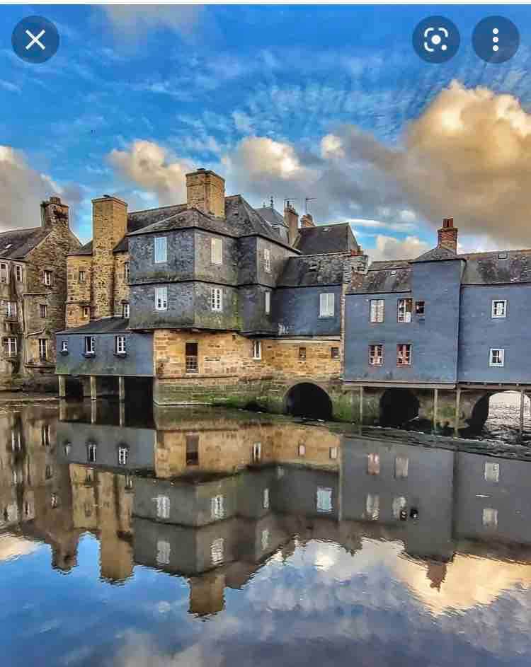 A collection of historic buildings featuring a mix of stone and blue facades is reflected in calm waters. The architecture showcases unique shapes and textures, with clouds softly mirrored above, creating a serene ambiance by the water's edge.