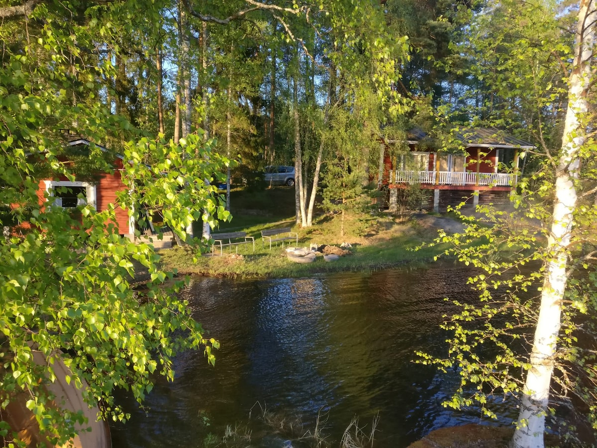 The image captures a traditional Finnish cottage surrounded by lush greenery, with sunlight filtering through leaves. A calm body of water reflects the environment. The structure features a wooden deck, enhancing outdoor access, while a rowboat and pier are visible along the shoreline.