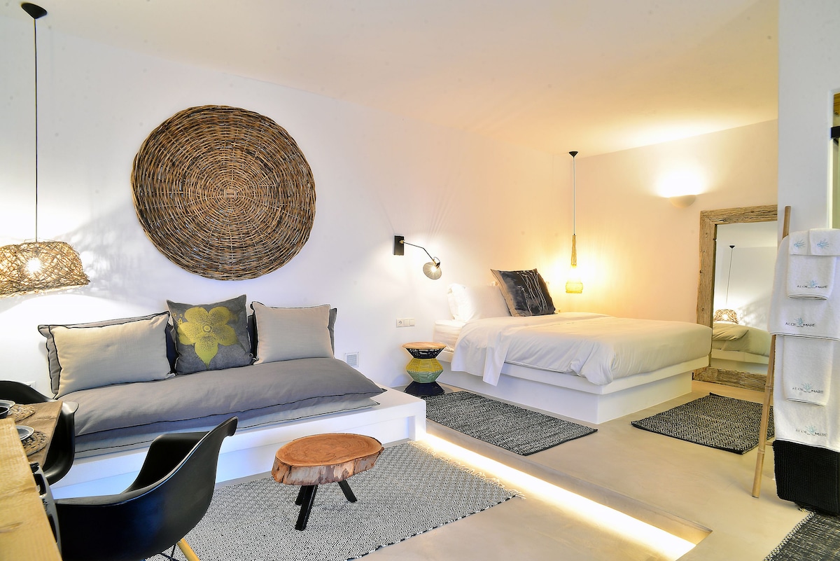 An inviting room showcases two spacious beds, one with crisp white bedding and the other adorned with decorative cushions. Natural textures are incorporated with a woven wall art piece and light wood accents. A wooden table and chairs provide a casual dining area.