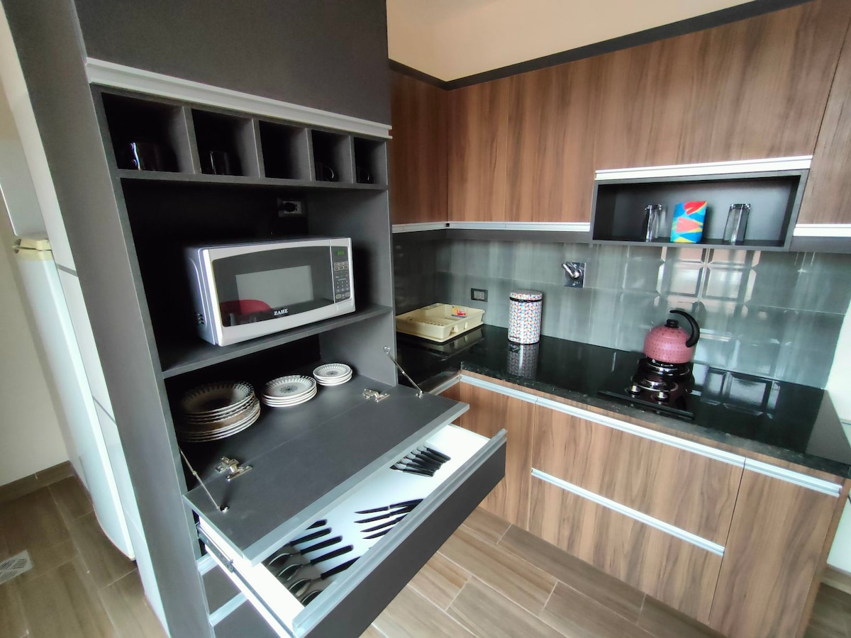 A modern kitchen area features wood cabinetry with a sleek black countertop. A microwave and kettle are visible on the counter. Drawers contain plates and utensils, indicating a well-organized space for meal preparation essentials.