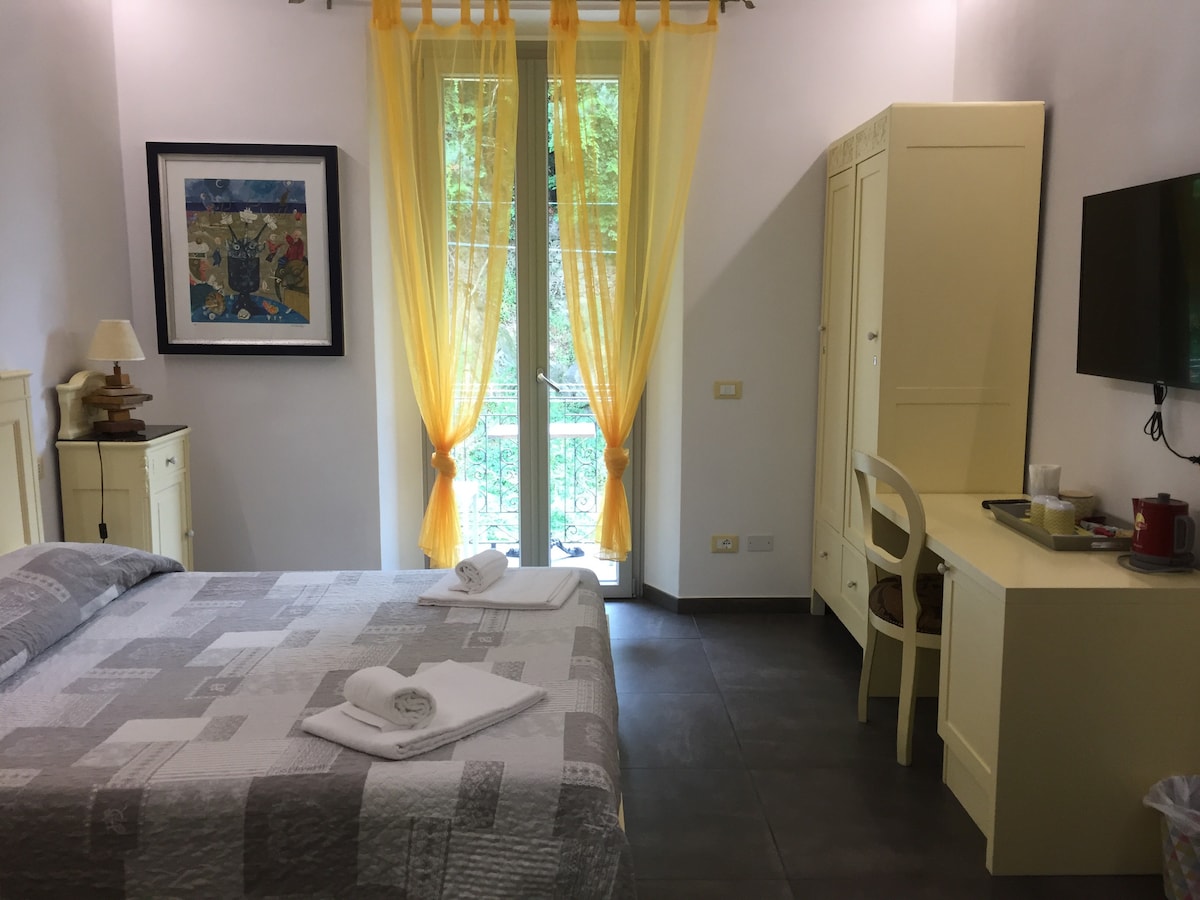 A well-lit room features a spacious bed covered with a light quilt. Yellow curtains frame a balcony door that opens to greenery. A wooden wardrobe and desk are positioned against the wall, with a flat-screen TV mounted above a small beverage station.