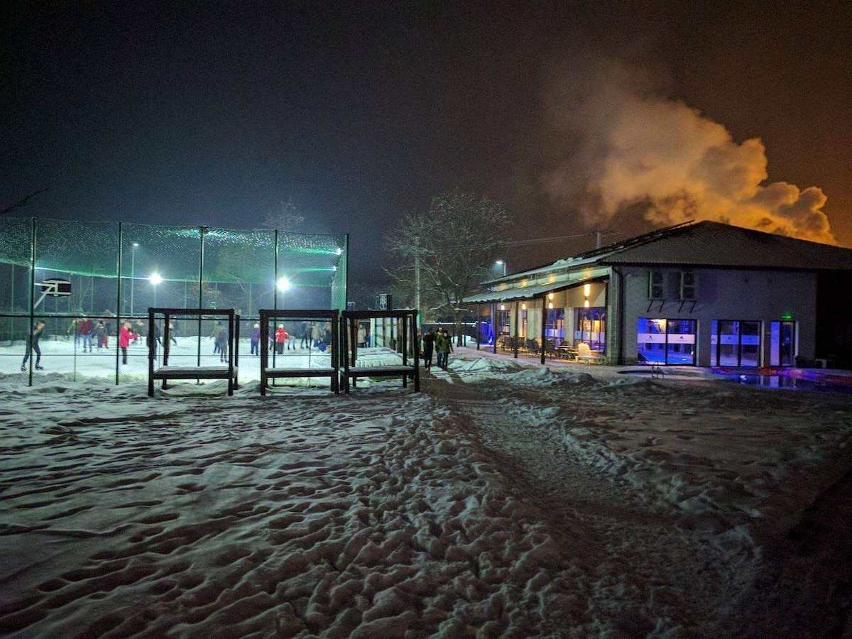 An outdoor ice skating rink is shown at night, surrounded by snowy terrain. Spotlights illuminate skaters who are enjoying the space, while a multi-sport court is visible nearby. A building with large windows is illuminated, creating a warm contrast against the cold landscape.