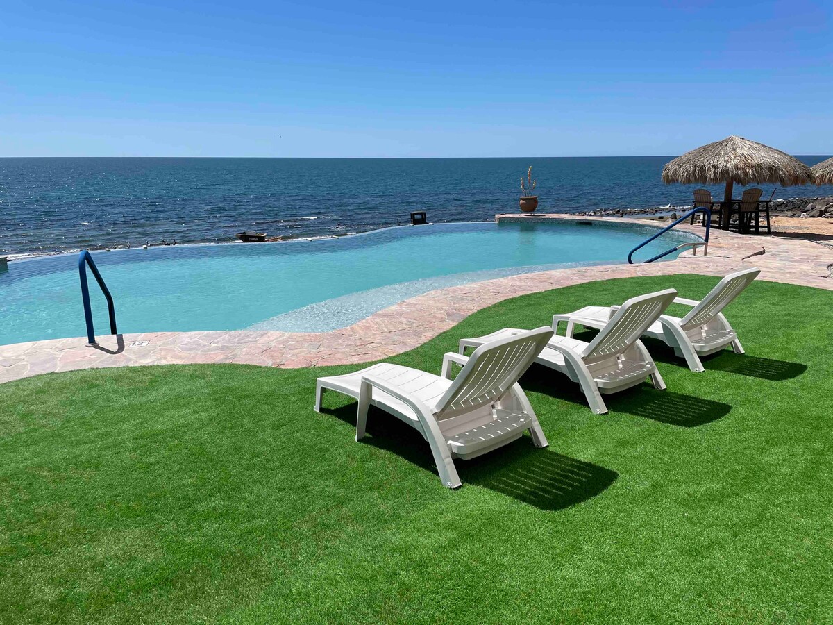 Lounge chairs are arranged by a serene pool that overlooks the ocean, framed by well-maintained grass. A thatched-roof umbrella provides shade nearby, while the expansive water and clear blue skies establish a relaxing atmosphere.