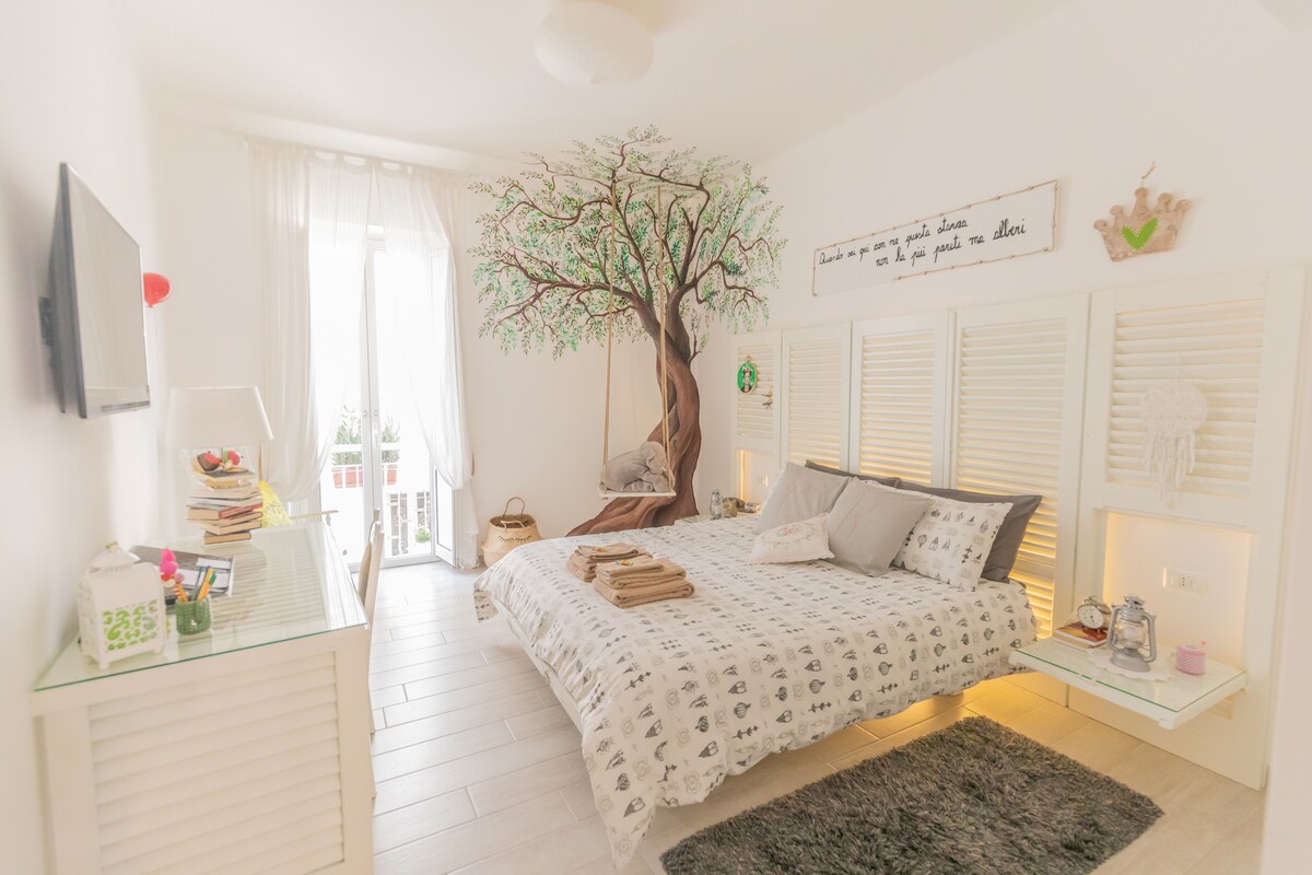 A bright bedroom features a spacious bed adorned with neutral bedding. A decorative tree mural adds a natural element, while a television is mounted on the wall. Natural light streams through sheer curtains, and a small desk is visible beside the bed.
