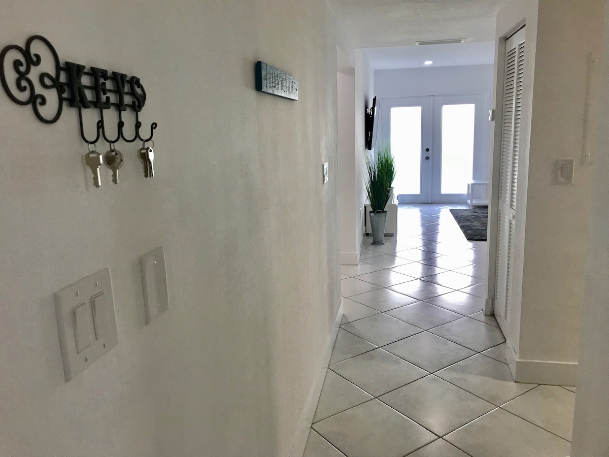 A bright entryway is shown, featuring a hallway with white walls and tiled flooring. A key rack hangs on the left side, and light filters through double glass doors at the end, leading to a spacious living area. A potted plant adds a touch of greenery.
