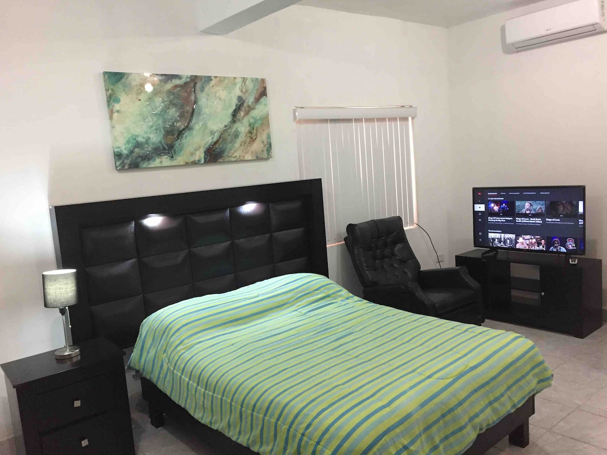 A comfortable sleeping area features a large bed with a striped green and blue blanket. A sleek black armchair sits nearby, accompanied by a small bedside lamp. A wall-mounted smart TV is displayed opposite the bed, providing modern entertainment options.