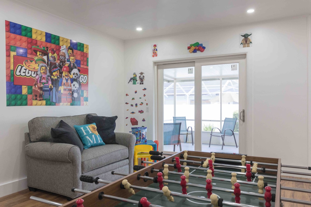 A playroom features a foosball table with red and yellow player figures, alongside a comfortable gray sofa adorned with a colorful cushion. Bright wall decals and Lego-themed decorations enhance the cheerful atmosphere, with large glass doors providing access to an outdoor seating area.