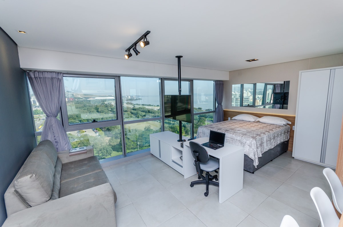 A spacious studio features a queen bed with a light-colored coverlet and a comfortable sofa. A desk with a chair is positioned next to a compact TV stand, providing a functional workspace. Large windows offer panoramic views of the surrounding greenery and distant water.