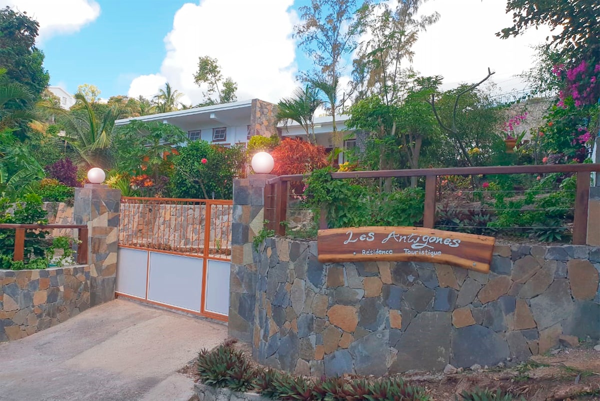 The entrance to Les Antigones is framed by a stone wall adorned with vibrant tropical plants. A wooden gate, marked with the name 'Les Antigones', leads into the property, which is highlighted by lush greenery and colorful flowers, creating a welcoming ambiance.