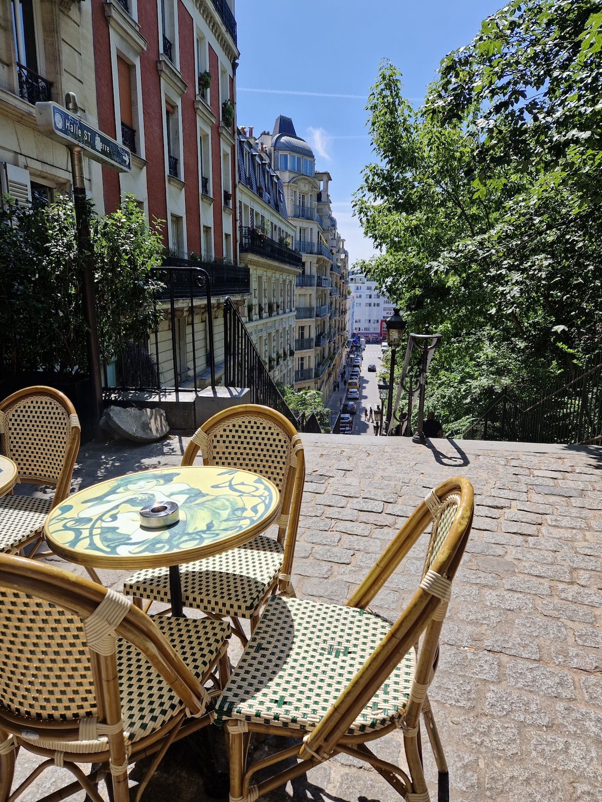 A charming outdoor seating area is visible, featuring a round table with a decorative top and four woven chairs. The surrounding environment includes lush greenery and a view of a tree-lined street, bathed in natural sunlight.