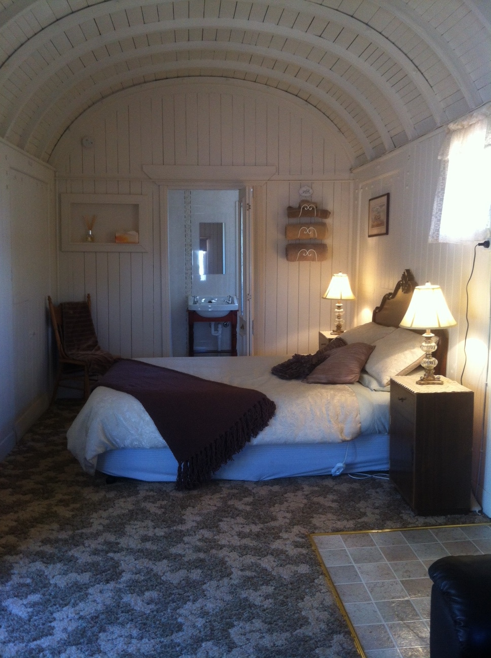 A quaint sleeping area features a spacious bed with soft linens and decorative pillows. Two bedside lamps provide warm lighting next to the bed. A small wooden table is positioned against the wall, while a mirror and additional decor are placed nearby. The arched wooden ceiling adds character.