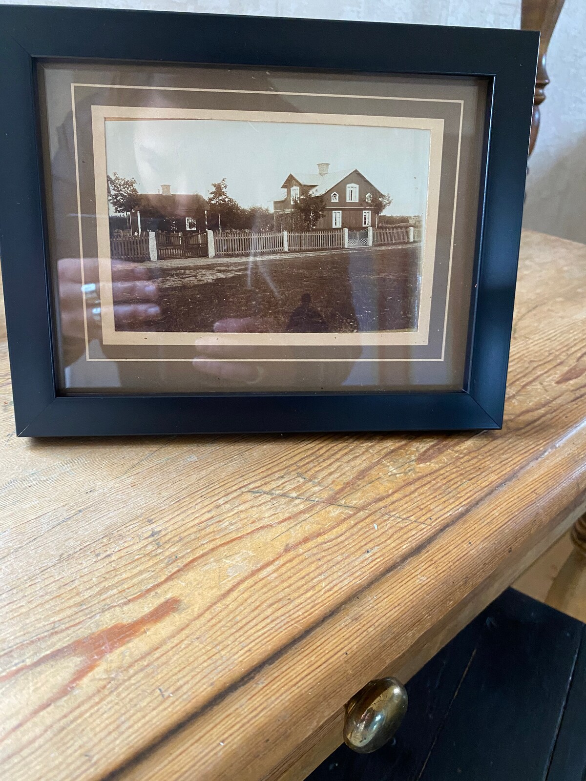 A framed black-and-white photograph sits on a wooden table. The image features a historic building with a fence in front, showcasing the architectural style of an earlier era. The frame is simple, contrasting with the warm tones of the wooden surface beneath.