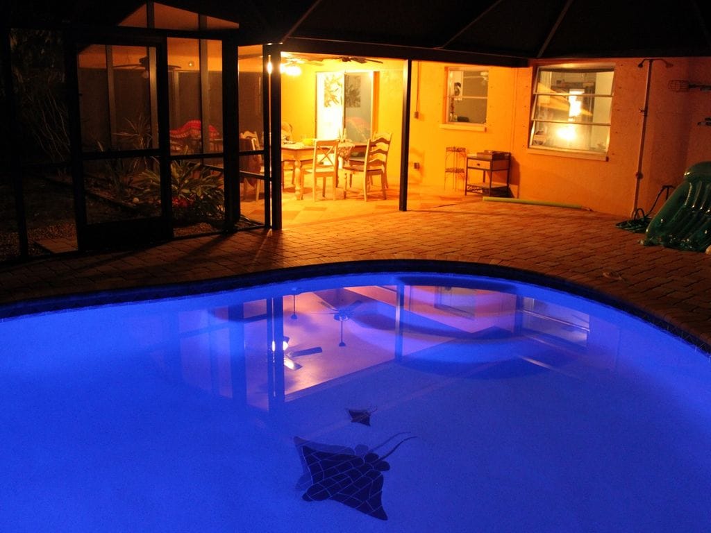 The image displays a heated caged swimming pool reflecting soft blue light. A dining area is visible in the background, illuminated by warm lighting, with a spacious table and chairs. The screened enclosure offers a view of the serene outdoor environment.