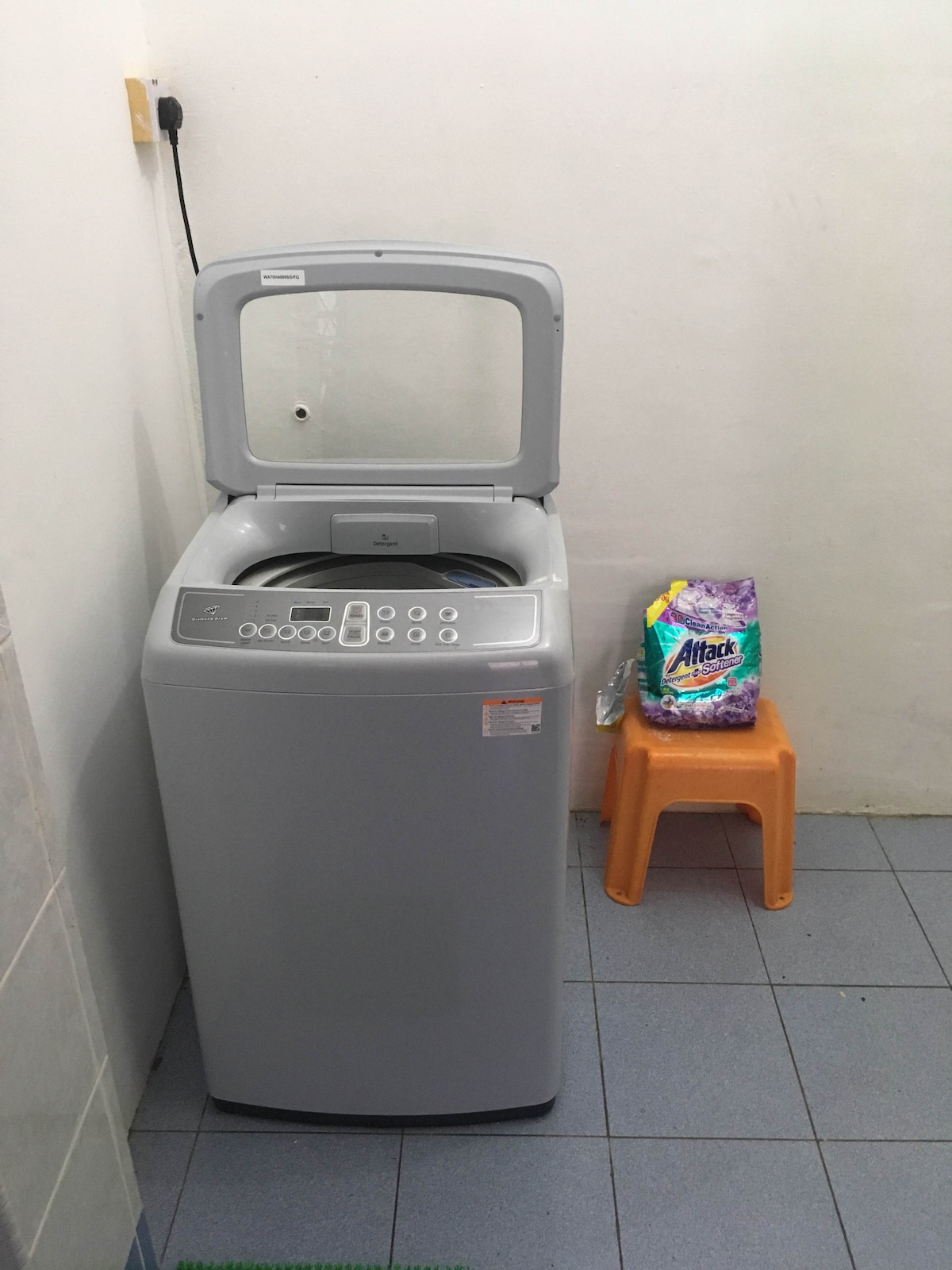 A washing machine is positioned against a plain wall, featuring a transparent lid and a control panel on the front. Next to it, a small orange stool holds a package of laundry detergent. The floor is tiled in a light blue color.