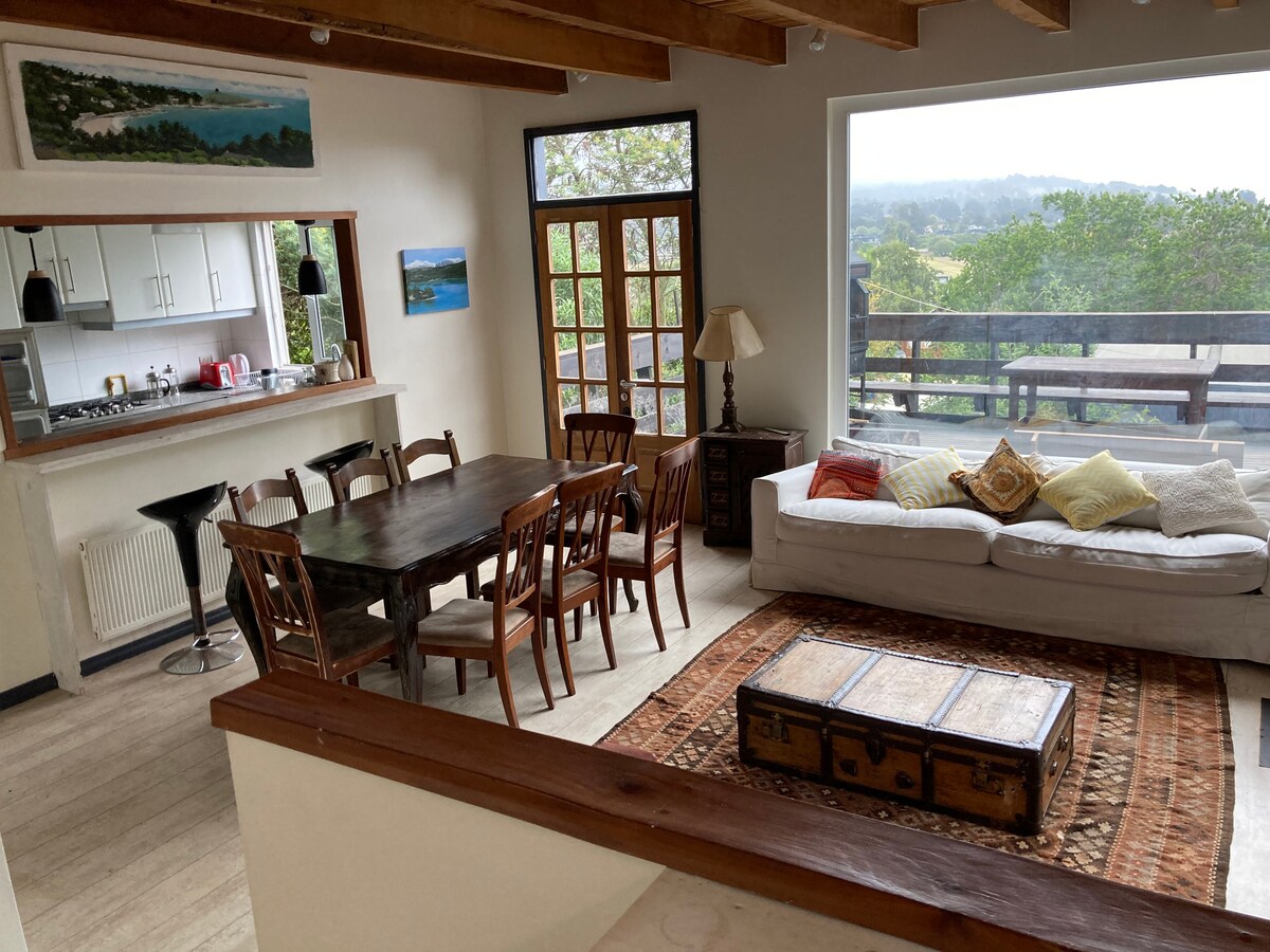 A spacious living area is equipped with a large dining table surrounded by wooden chairs, and a comfortable sofa adorned with pillows. A coffee table sits in front of the sofa on an area rug, while large windows offer views of the outside greenery.