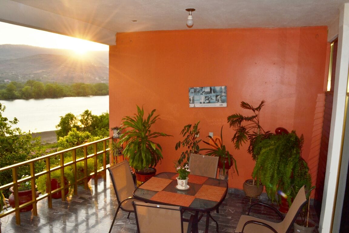 A terrace area features a table surrounded by four chairs and potted plants. The wall is painted a warm orange, and a scenic view of the river is visible in the background as the sun sets.