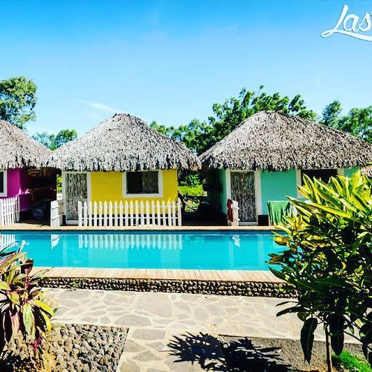 A tranquil scene features colorful thatched-roof cabins surrounding a clear, inviting pool. Lush greenery and tropical plants are planted along the stone pathway leading to the cabins. The vibrant colors of the cabins contrast beautifully against the blue sky.