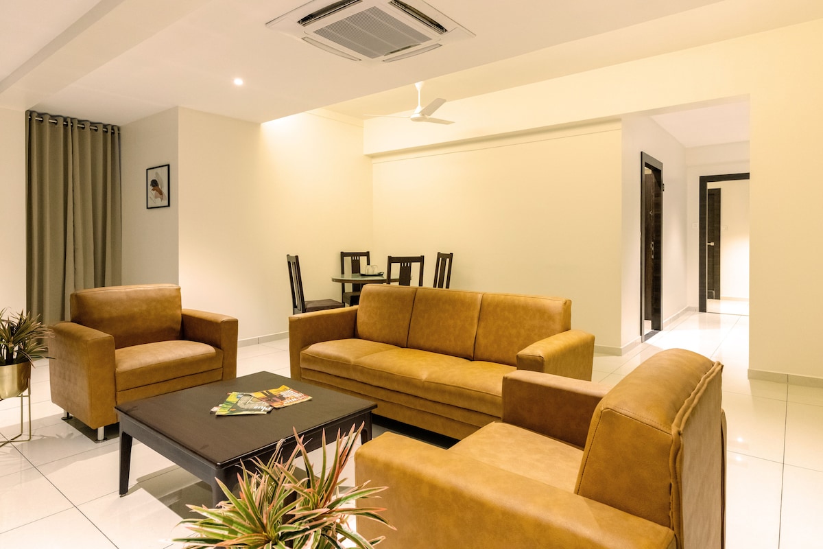 A spacious living area features two tan leather sofas and a dark coffee table. A small dining set is visible in the background, complemented by modern decor. Light-colored walls and polished tiles enhance the room's openness, while indoor plants add a touch of greenery.