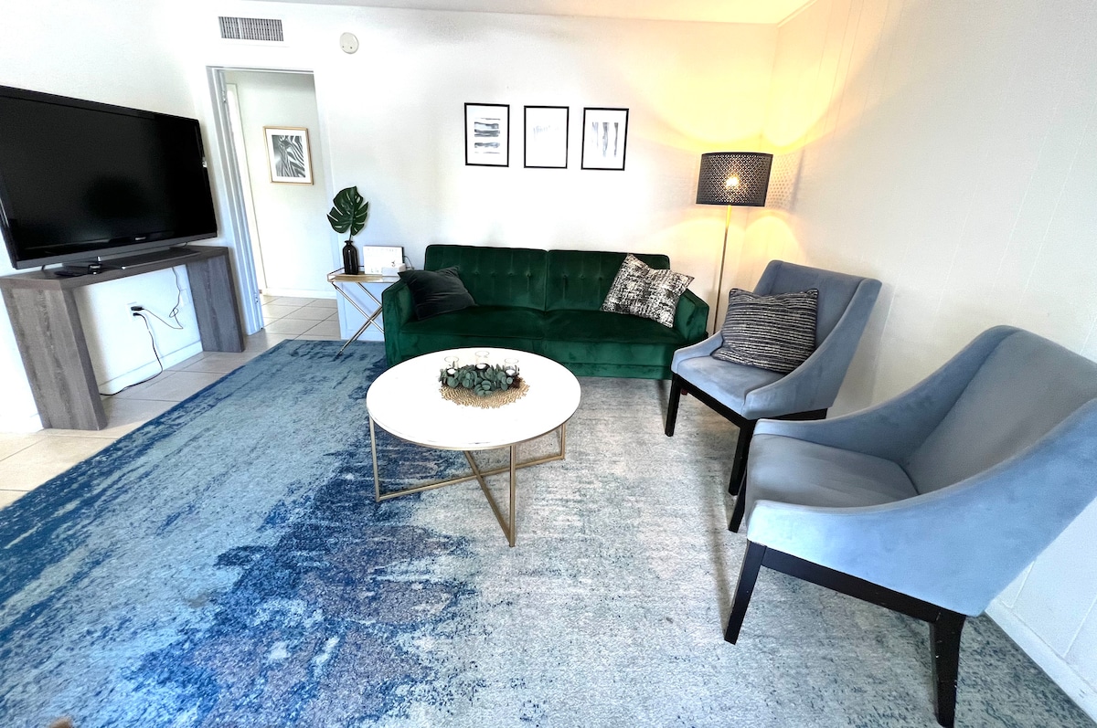 A spacious living area features a green couch and two accent chairs positioned around a circular coffee table. A flat-screen television is mounted on the wall, and natural light illuminates the room through a nearby doorway. A textured rug adds softness underfoot.