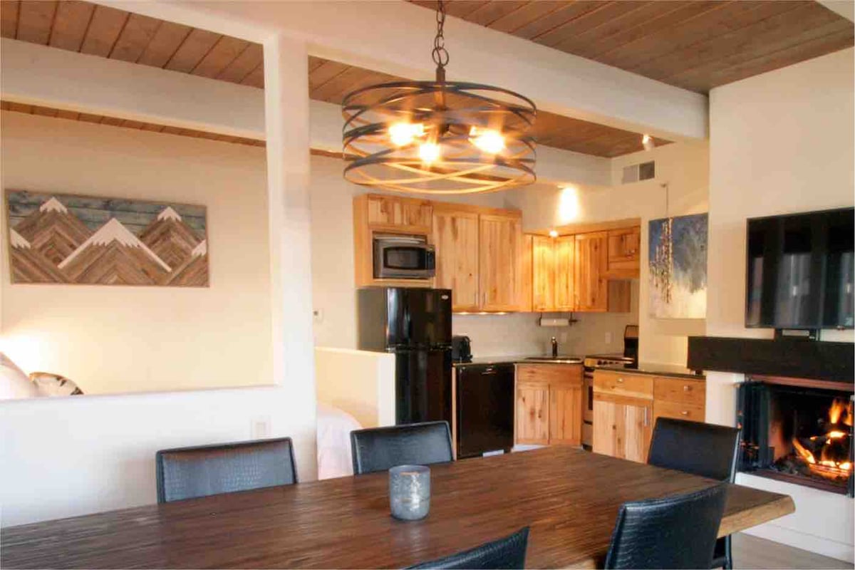An open dining area features a warm wooden table surrounded by black wicker chairs. Above, a circular light fixture adds a modern touch. The kitchen is visible with wooden cabinetry, appliances, and a black refrigerator, while a gas fireplace glows softly in the corner.