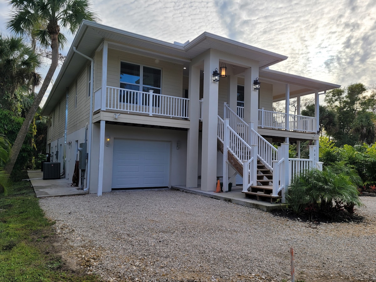 The exterior of a modern two-story home is featured, showcasing a spacious garage at ground level. Sturdy steps lead to a wide porch with column supports and railing. Lush greenery surrounds the property, with palm trees and a gravel driveway adding to the tranquil setting.