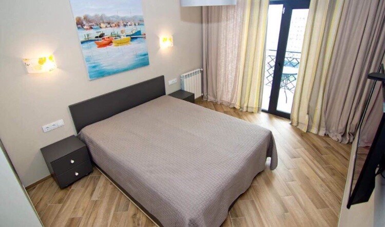 A light-filled bedroom features a spacious bed covered with a neutral-toned blanket. Two bedside tables flank the bed. A large window is adorned with light, flowing curtains, which open to a view outside. A piece of wall art depicting boats adds a touch of color.