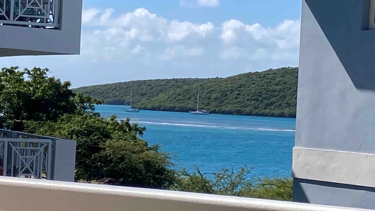 A view of the serene bay is framed by lush green hills and a clear blue sky. Two sailboats are anchored in the calm waters, while gentle waves can be seen near the shoreline. The scene conveys a peaceful coastal atmosphere.