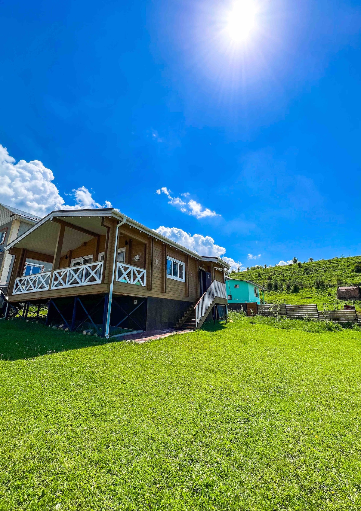The house is positioned on a green hillside, featuring a shaded porch with white railings. Surrounding areas include grassy fields and distant hills under a clear blue sky with scattered clouds.