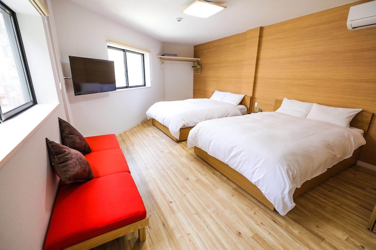The room contains two spacious beds dressed in crisp white linens. A red seating area is positioned beside the beds. A wall-mounted television is visible, and natural light enters through the window, highlighting the warm wooden tones of the decor.