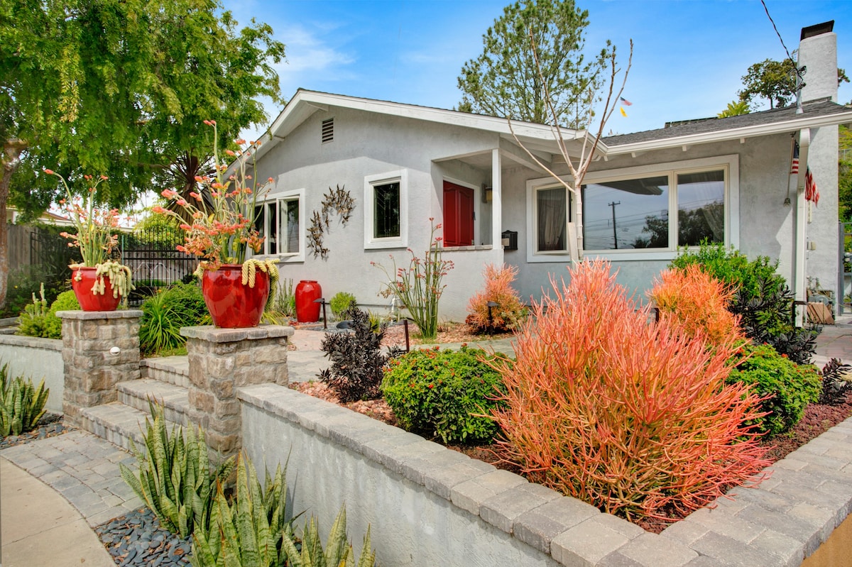 A well-maintained exterior features colorful landscaping with vibrant plants and flowers. Decorative pots in red add pops of color against the neutral gray facade of the house. A stone pathway leads to the entrance, flanked by lush greenery and a clear blue sky above.
